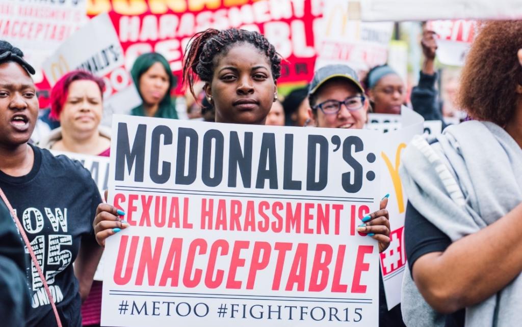 A Black woman at a demonstration faces the camera holding a sign that reads "McDonald's: sexual harassment is unacceptable #MeToo #FightFor15. She has black braids with a red streaks, tied up. Credit: FightFor15 Chicago