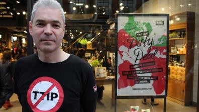 Executive Director John Hilary, wearing a No TTIP t shirt, stands in front of a Lush store that has an advert in the window reading: "Vivienne Westwood; TTIP that dirty deal down the plughole; This is a people-power shower; don't let TTIP wash away our rights....; waronwant.org/ttip"