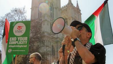 A protestor speaking into a megaphone in front of the Palestinian flag. A placard to the left reads "HSBC: End Your Complicity".